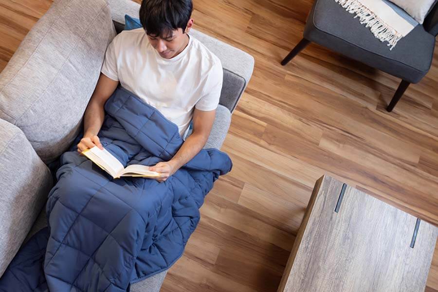 Man reading a book on the couch, with Classic Weighted Blanket draped over his lower body